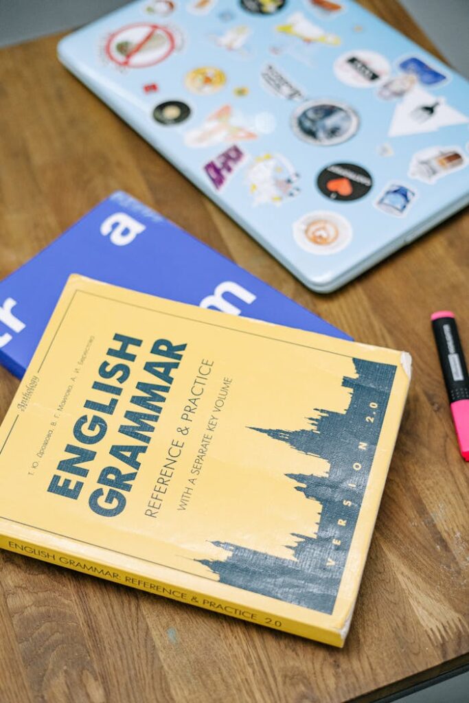 pexels-photo-5676740 High-angle view of an English grammar book and a laptop with stickers on a wooden desk.